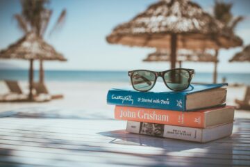Pila de libros y gafas de sol sobre una mesa en la playa, representando la preparación flexible de exámenes oficiales de inglés B2 y C1 durante las vacaciones de verano