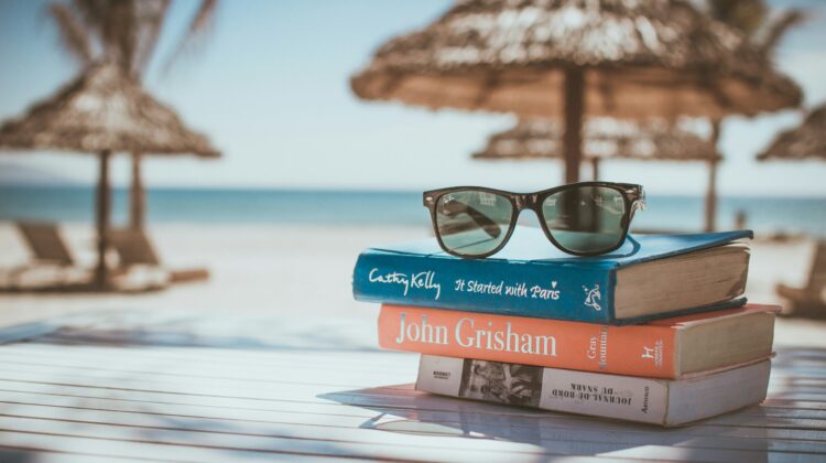 Pila de libros y gafas de sol sobre una mesa en la playa, representando la preparación flexible de exámenes oficiales de inglés B2 y C1 durante las vacaciones de verano