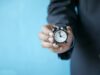Primer plano de una mano con traje sosteniendo un pequeño reloj despertador clásico, representando la caducidad y los límites de tiempo de los certificados de inglés en las oposiciones
