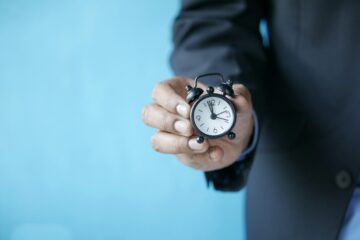 Primer plano de una mano con traje sosteniendo un pequeño reloj despertador clásico, representando la caducidad y los límites de tiempo de los certificados de inglés en las oposiciones