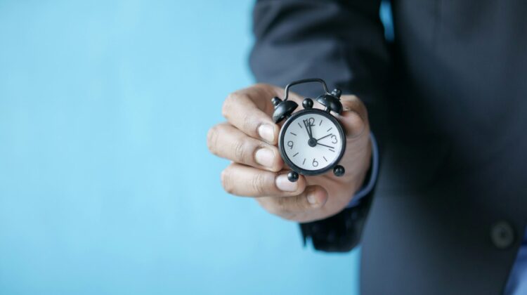 Primer plano de una mano con traje sosteniendo un pequeño reloj despertador clásico, representando la caducidad y los límites de tiempo de los certificados de inglés en las oposiciones