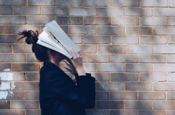 Alumna abrumada cubriéndose la cara con un libro abierto mientras se apoya en una pared de ladrillo, representando la frustración de preparar exámenes oficiales de inglés B2 y C1 sin el método adecuado