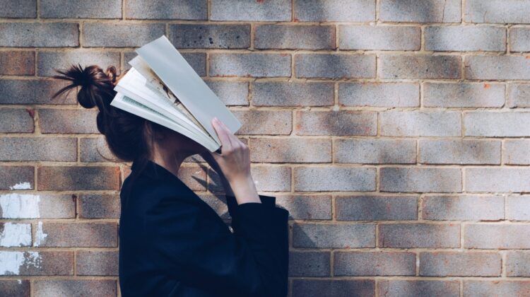 Alumna abrumada cubriéndose la cara con un libro abierto mientras se apoya en una pared de ladrillo, representando la frustración de preparar exámenes oficiales de inglés B2 y C1 sin el método adecuado