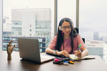 Alumna con auriculares tomando notas frente a un ordenador portátil, simulando la preparación de la prueba de comprensión auditiva y oral de inglés