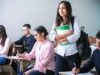 Grupo de estudiantes universitarios en un aula, con una alumna de pie sonriendo con su mochila y libros, representando la obtención del certificado de inglés para el grado