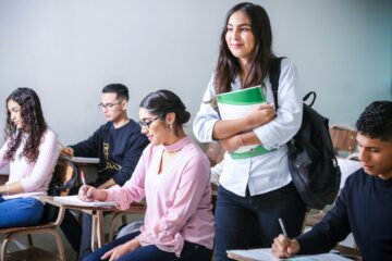 Grupo de estudiantes universitarios en un aula, con una alumna de pie sonriendo con su mochila y libros, representando la obtención del certificado de inglés para el grado
