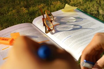 Primer plano de un estudiante usando un subrayador naranja sobre un libro de texto abierto en el césped, con unas gafas apoyadas en las páginas, representando el estudio al aire libre durante el verano