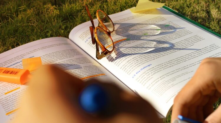 Primer plano de un estudiante usando un subrayador naranja sobre un libro de texto abierto en el césped, con unas gafas apoyadas en las páginas, representando el estudio al aire libre durante el verano