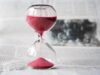Reloj de arena clásico de cristal con arena rojiza sobre un periódico, simbolizando el tiempo de estudio necesario para preparar certificaciones oficiales de inglés