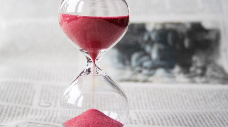 Reloj de arena clásico de cristal con arena rojiza sobre un periódico, simbolizando el tiempo de estudio necesario para preparar certificaciones oficiales de inglés