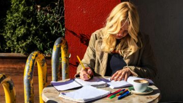 Mujer joven escribiendo relajadamente en una libreta mientras toma un café en una terraza soleada, representando la actitud natural necesaria para redactar un email informal en inglés