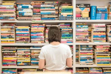 Mujer de espaldas mirando una enorme estantería llena de libros y diccionarios, representando la frustración de intentar memorizar interminables listas de Phrasal Verbs en inglés