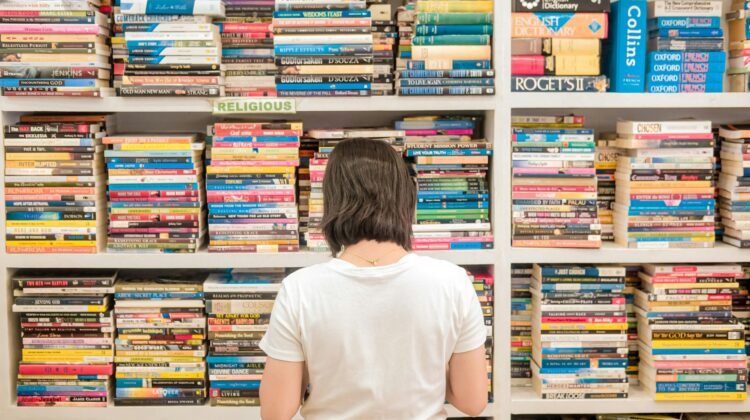 Mujer de espaldas mirando una enorme estantería llena de libros y diccionarios, representando la frustración de intentar memorizar interminables listas de Phrasal Verbs en inglés