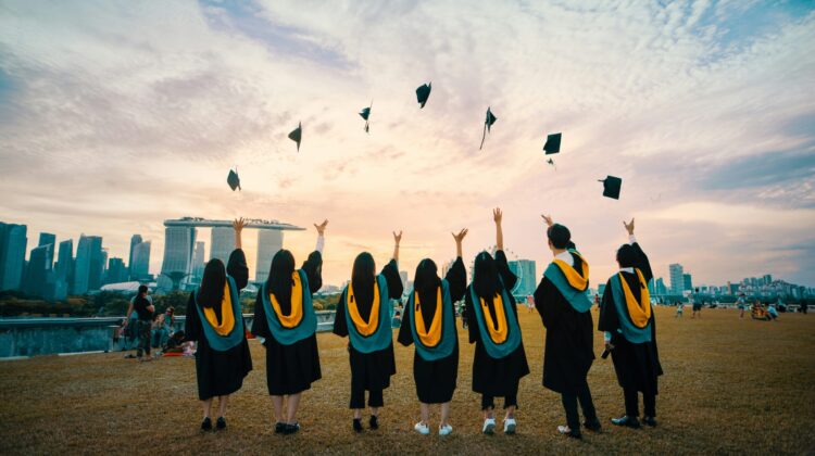 Grupo de estudiantes universitarios vestidos con toga, de espaldas, lanzando sus birretes al aire durante el atardecer para celebrar el fin de su carrera