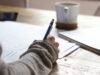 Primer plano del brazo de un estudiante escribiendo con bolígrafo sobre unos apuntes en una mesa de madera, representando la planificación de un ensayo de opinión en inglés