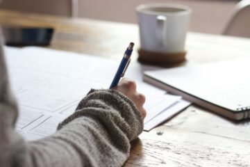 Primer plano del brazo de un estudiante escribiendo con bolígrafo sobre unos apuntes en una mesa de madera, representando la planificación de un ensayo de opinión en inglés