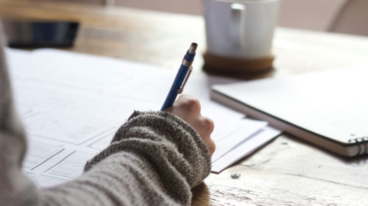 Primer plano del brazo de un estudiante escribiendo con bolígrafo sobre unos apuntes en una mesa de madera, representando la planificación de un ensayo de opinión en inglés