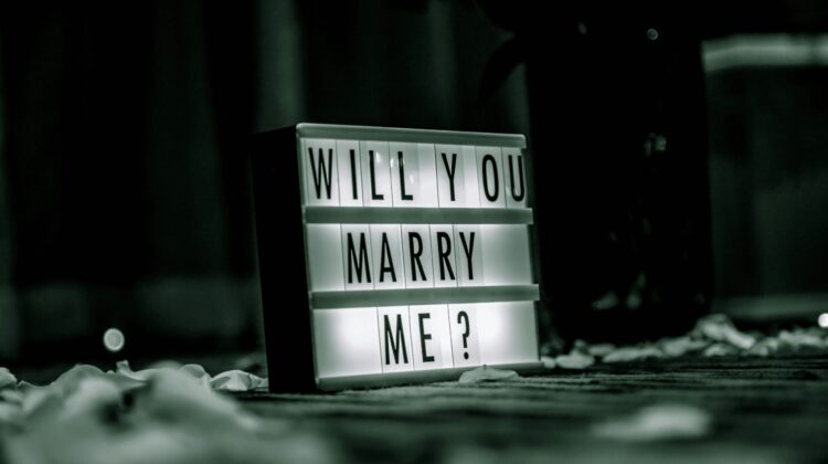 Cartel luminoso con el texto Will you marry me rodeado de pétalos, representando el juego de palabras del término Proposal en inglés
