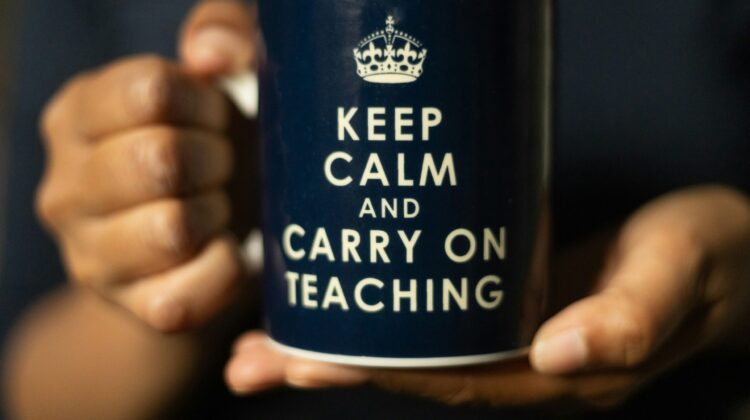 Persona sujetando con ambas manos una taza azul oscuro con el mensaje en inglés Keep Calm and Carry on Teaching