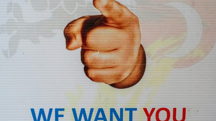 Cartel con una mano que señala al espectador y el texto «WE WANT YOU» en letras azules y rojas sobre fondo blanco