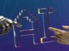 Mano robótica metálica y mano humana extendiendo el dedo hacia las letras «AI» de cristal sobre un fondo azul con motivos de circuito electrónico